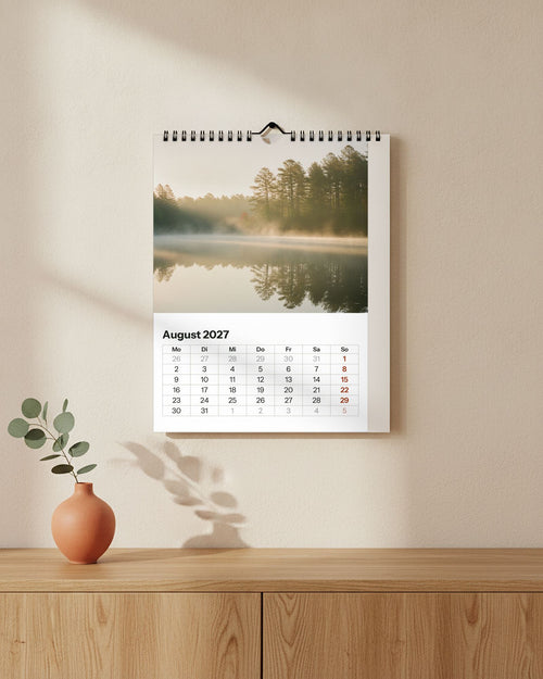 Großer Wandkalender hochkant an cremefarbener Wand über einem Sideboard: Seefoto im Morgennebel, darunter das Monatsraster August 2027